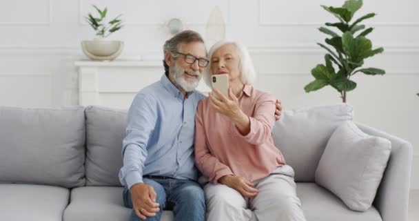 Biały starszy mężczyzna i kobieta na emeryturze. Szczęśliwa para siedząca na kanapie i pozująca z uśmiechem do kamery telefonicznej w domu. Stara para robi zdjęcia ze smartfonem. - Materiał filmowy, wideo