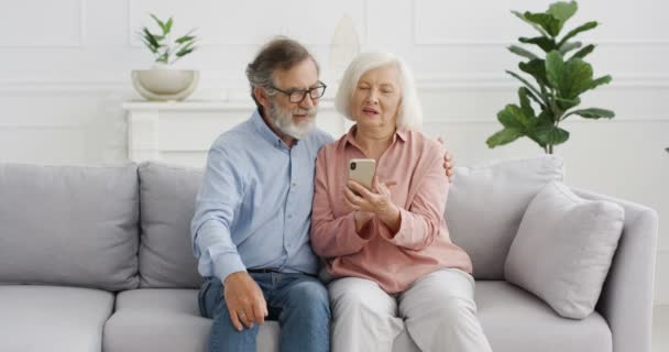 Oude blanke vrolijke paar zitten op de bank en kijken naar iets op smartphone scherm en praten. Thuis. Oudere gepensioneerde man en vrouw met behulp van mobiele telefoon, afluisteren en scrollen, bespreken. - Video
