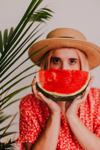 Young beautiful lady in red dress and straw hat is holding juicy watermelon. Red bright color. Green tropical. Summer time vacation. Beach vibes. Hot days. Refreshing red fruit.Pretty millennial woman - Photo, Image