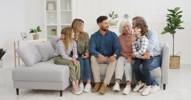 大きな白人の家族がリビングルームでソファに座ってコミュニケーションを取っています。ロックダウン時に自宅で祖父母、両親と小さな子供。母親、父親、祖母、祖父と話す子供たち. - 映像、動画