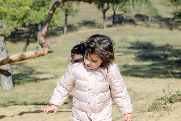 Biała dziewczyna przedszkolak z trzepoczącymi włosami w ciepłej kurtce wznosi się pod górę w lesie na tle drzew i trawy - Zdjęcie, obraz