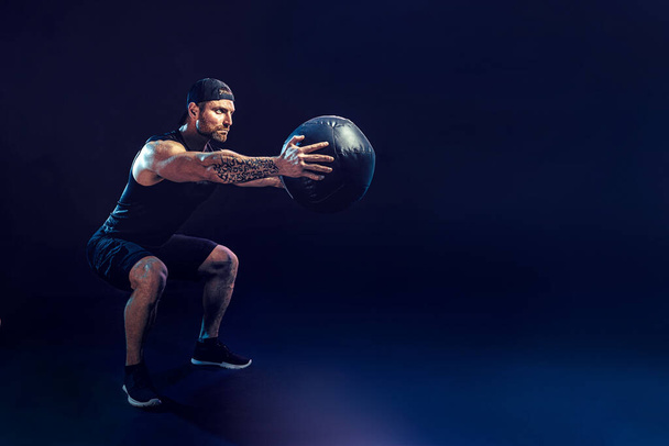 Aggressive bearded muscular sportsman is working out with a medicine ball isolated on dark studio background - Foto, Bild