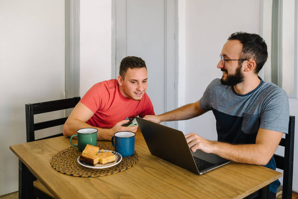 Photo de deux hommes adultes caucasiens assis à une table et souriant. L'un d'eux travaille avec un ordinateur portable et l'autre utilise son smartphone. - Photo, image