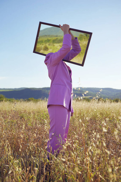 Side view portrait full length of unknown woman standing in the field hold mirror on shoulder covering head - Female hiding identity standing out from the crowd concept - Female person lost in nature - Photo, Image
