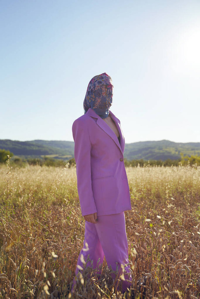 Boční pohled portrét po celé délce na neznámé ženě v purpurovém obleku stojící na poli v přírodě ve slunečný letní den nebo večer se šátkem na hlavě - identita a matka příroda zmatek koncept - Fotografie, Obrázek