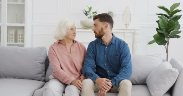 Caucasian young handsome man sitting on couch and talking nice with old beautiful mother. Senior gray-haired woman pensioner embracing and talk with adult son. At home. Motherhood love and support. - Footage, Video