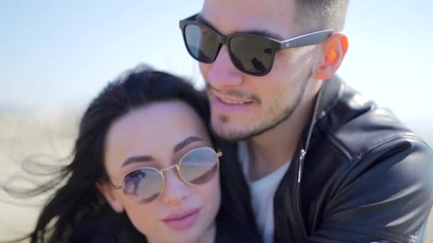 Close up portrait of a stylish fashion young couple in black leather jackets and sunglasses. A man hugs a woman from behind. black hair. Caucasians. Attractive and beautiful. Luxury guy and girl - Кадри, відео