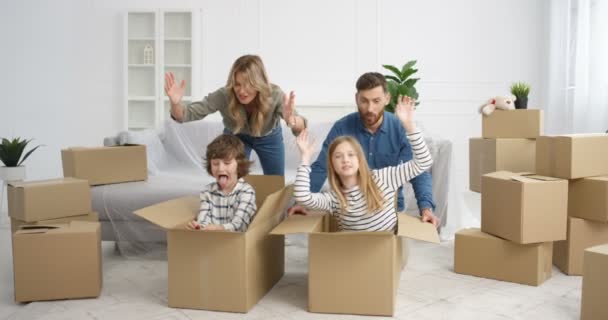 Nuoret valkoihoiset vanhemmat pitävät hauskaa kotona lasten kanssa muuttaessaan uuteen taloon. Söpö pieniä lapsia, poika ja tyttö istuu pahvilaatikoissa ja nauraa leikkiessään äidin ja isän kanssa asunnossa - Materiaali, video