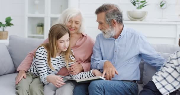 Velhos avós caucasianos com netos sentados no sofá em casa e lendo livro. Avó, avô, neto e neta passando tempo juntos e estudando com livro didático. Interior. - Filmagem, Vídeo