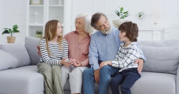 Kavkazští staří prarodiče s vnoučaty sedí na gauči v útulném obývacím pokoji. Babička, dědeček, vnuk a vnučka spolu tráví čas doma. Dovnitř. Rodinné generace. - Záběry, video
