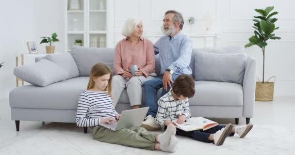 Aranyos kis kaukázusi gyerekek játszanak a nappaliban. Nagymama és nagyapa a kanapén ülnek és a háttérben beszélgetnek. A kisfiú könyvet olvas és laptopot használ. Otthon. Együtt a család. - Felvétel, videó