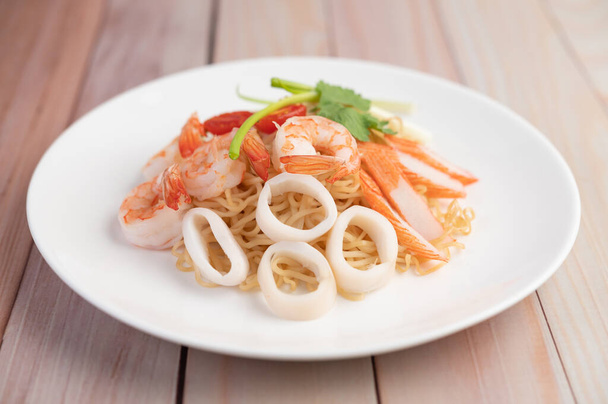 Stir-fried instant noodle with prawn and crab stick along with tomatoes and peppers in a white plate on a wooden floor. - Photo, Image