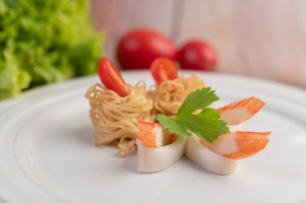 Stir-fried instant noodle with prawn and crab stick along with tomatoes and peppers in a white plate on a wooden floor. - Photo, Image
