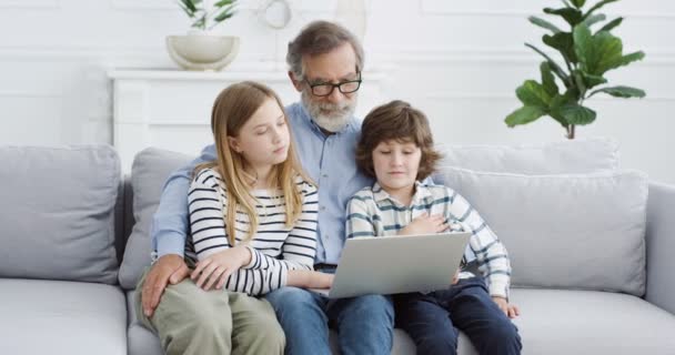 avô caucasiano sentado no sofá com duas crianças pequenas e conversando. Avô abraçando crianças pequenas no sofá e usando juntos computador portátil. Em casa. Homem sênior em óculos com netos. - Filmagem, Vídeo