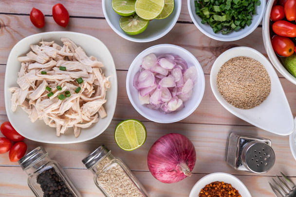 Boiled chicken cut into pieces in a white dish, along with lemons, chilies, lemongrass, red onions, chopped onions, tomatoes, sesame, roasted rice, and pepper on a wooden table. - Photo, Image