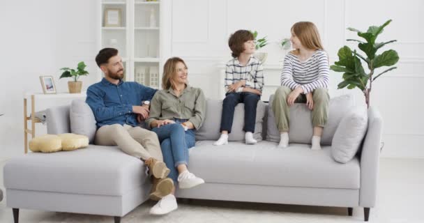 Aranyos kis fehér gyerekek és szülők ülnek a kanapén a nappaliban és beszélgetnek. Anya és apa a kanapén otthon és kommunikálnak. A család együtt töltött egy kis időt az elszigetelt zárlatban.. - Felvétel, videó
