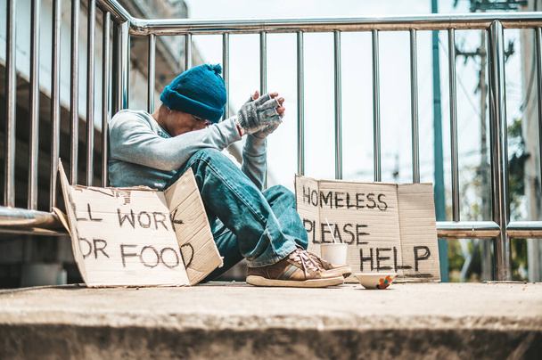 Человек, сидящий попрошайничая на эстакаде с сообщениями бездомные люди, пожалуйста, помогите и хорошо работать с едой. - Фото, изображение