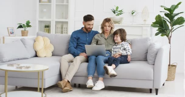 若い白人の両親はソファに座って、ラップトップを使用して小さなかわいい息子を教えています。小さな男の子とお母さんとお父さんのコンピュータ上で何かを見てリビングルームで. - 映像、動画