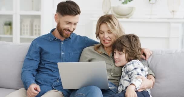 Happy small cute child son sitting in hugs with parents on couch and shopping online with credit card on laptop computer. Mother father with little kid boy buying in internet and paying. - Footage, Video