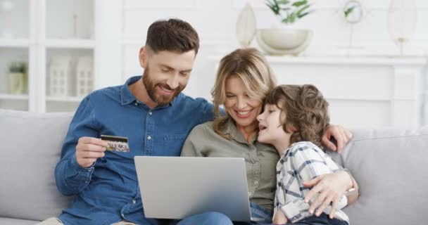 Caucasian family with child spending money together on sofa at home. Parents with son using laptop to buy online with credit card. Kid with mother and father shopping in internet on computer. - Footage, Video