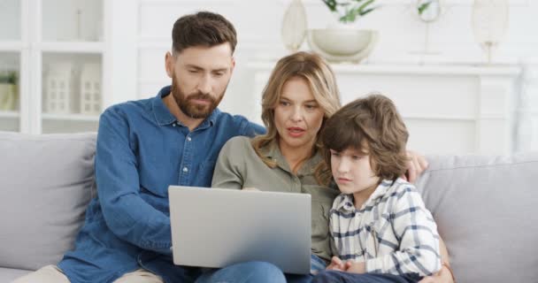 Καυκάσιοι νεαροί γονείς με μικρό παιδί να κάθεται στον καναπέ στο σπίτι, χτυπώντας και κυλώντας σε φορητή συσκευή. Μητέρα, πατέρας διδασκαλία μικρό γιο παίζει και χρησιμοποιώντας υπολογιστή και δείχνει βίντεο Οικογένεια μαζί - Πλάνα, βίντεο