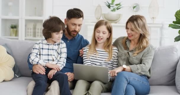 Iloiset lapset istuvat halauksissa vanhempien kanssa sohvalla ja katsovat videota kannettavasta tietokoneesta. Äiti isä pieniä lapsia, tytär ja poika puhuu ja vierittää verkossa Perhe on lukitus käsite - Materiaali, video