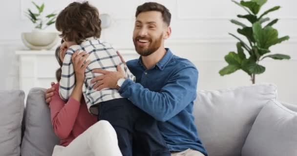 Caucasian cute little kid boy running to young happy mother and father sitting on couch in cozy living room. Cheerful parents hugging and embracing their small son at home on sofa. Indoors. - Кадры, видео