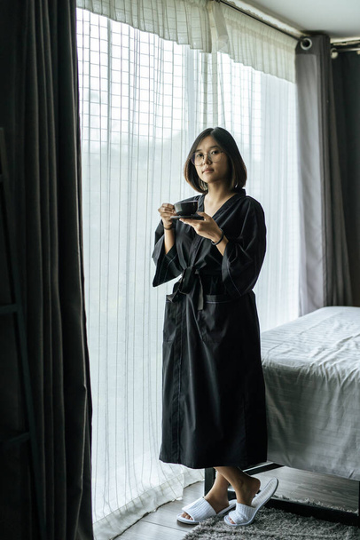 The woman wore a black robe near the window and drank coffee in the bedroom. - Photo, Image