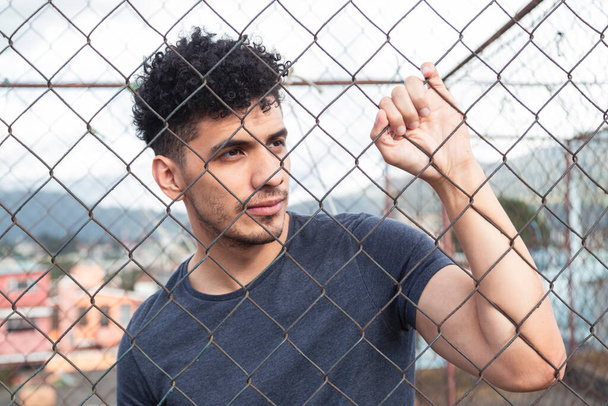 Retrato de un joven hispano frente a una malla metálica al atardecer - un joven pensativo que reflexiona en la azotea - estilo urbano
 - Foto, Imagen