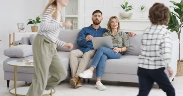 Two cheerful Caucasian kids running in circles and rounds in living room. Happy cute small sister and brother playing around sofa. Smiled parents sitting on couch with laptop computer Hugging children - Footage, Video