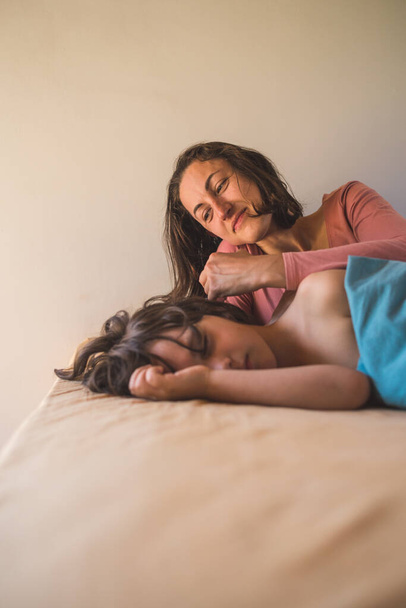 Η μητέρα χαϊδεύει το κεφάλι ενός παιδιού που κοιμάται, μια γυναίκα αγγίζει τα μαλλιά του γιου της, το αγόρι αποκοιμιέται στο κρεβάτι κοντά στη μητέρα του.. - Φωτογραφία, εικόνα