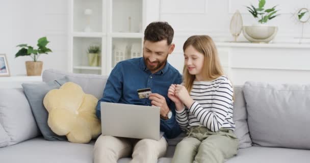 Valkoihoinen isä istuu sohvalla huoneessa onnellinen tytär käyttää kannettavaa tietokonetta ja ostoksia verkossa luottokortilla. Pikkutyttö ostaa internetin isän kanssa. Sisätiloissa. Lapsi shopper rahaa isän kanssa - Materiaali, video