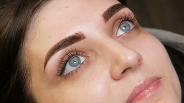 Das Gesichtsporträt einer schönen jungen Frau mit blauen Augen und langen Wimpern nach der Prozedur des Permanent Make-up durch Microblading mit Augenbrauentätowierung liegt auf einer Couch in einem Schönheitssalon - Filmmaterial, Video