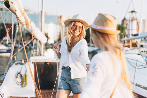 Two beautiful blonde girls friends mother and daughter in white and straw hats on the yacht at pier - 写真・画像