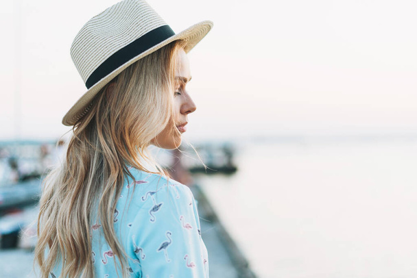 Belle jeune femme blonde en robe bleue et chapeau de paille sur jetée au coucher du soleil - Photo, image