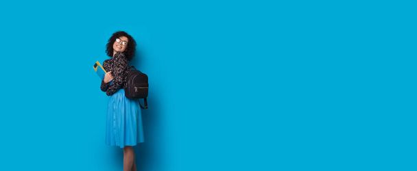 Curly haired student wearing eyeglasses and bag holding some folders is smiling on a blue studio wall with free space - Photo, Image