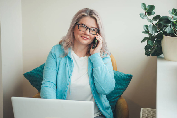 Idős fehér nő szőke hajjal és szemüveggel telefonon beszél és távirányítással dolgozik a laptopnál. - Fotó, kép