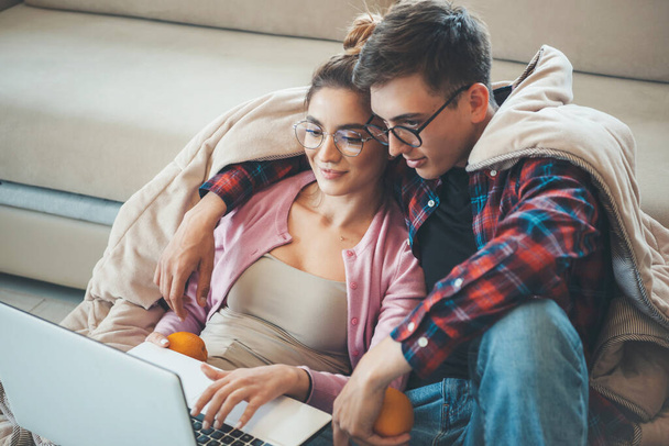 Young caucasian couple sitting on floor covered with a quilt holding an orange and looking at the laptop - Foto, Imagem