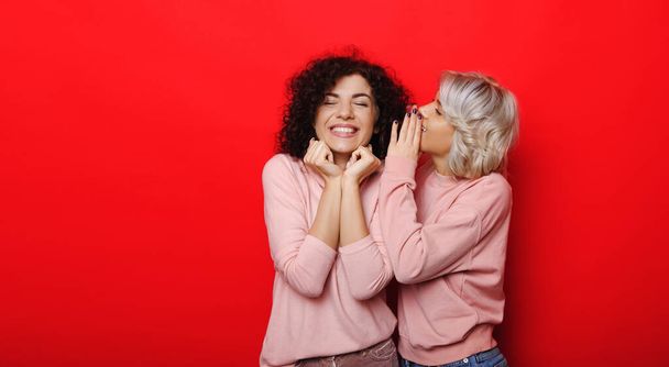Curly haired blonde woman whispering something to her brunette friend dreaming on a red studio wall - Photo, Image