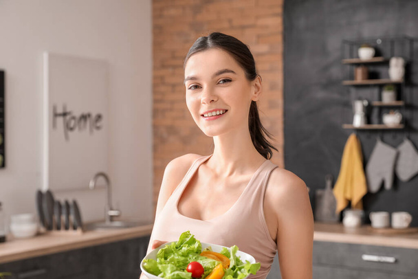 Beautiful young woman eating vegetable salad in kitchen - Fotó, kép