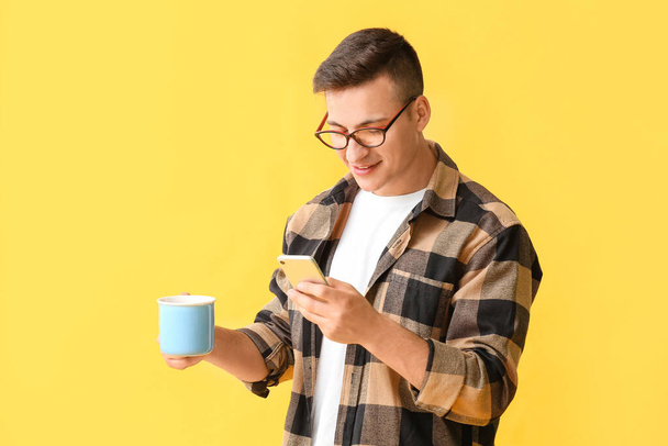Handsome young man with cup of tea and mobile phone on color background - Photo, Image