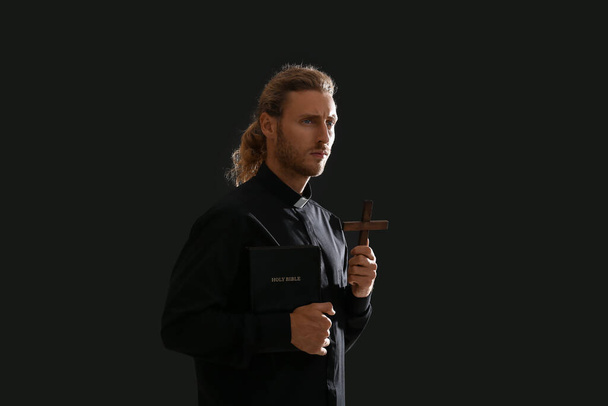 Όμορφος ιερέας με Βίβλο και σταυρό σε σκούρο φόντο - Φωτογραφία, εικόνα