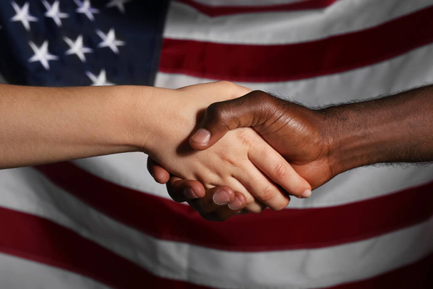 Καυκάσια γυναίκα και Αφροαμερικάνος κάνουν χειραψία κοντά στην εθνική σημαία των ΗΠΑ. Έννοια ρατσισμού - Φωτογραφία, εικόνα