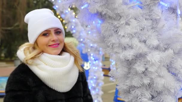 Sıcak şapkalı mutlu kadın, atkı beyaz Noel ağacının yanında duruyor, gülümsüyor, poz veriyor. - Video, Çekim