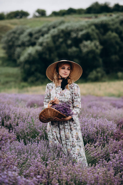 Young beautiful blonde woman in romantic dress, straw hat and basket of flowers posing in lavender field. Soft selective focus. - Fotografie, Obrázek