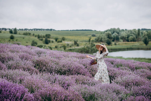 Young beautiful blonde woman in a romantic dress, a straw hat and a basket of flowers dancing in a lavender field. Soft selective focus. - Foto, Bild