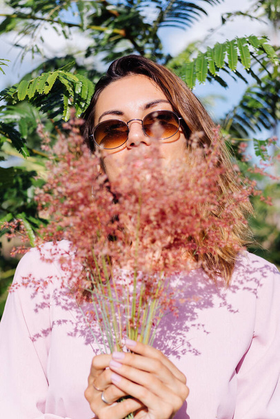Retrato de jovem caucasiana bronzeada em vestido rosa romântico, brincos redondos, pulseira de prata e óculos de sol, com flores silvestres, em fundo tropical exótico. Mulher bonita calma desfrutando do sol no parque
 - Foto, Imagem