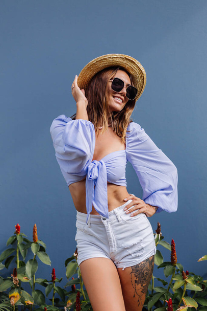 Pretty young stylish woman in trendy purple crop top long sleeve , white shorts, black sunglasses, straw hat and pearl necklace, isolated, blue gray wall, natural day light. - Valokuva, kuva