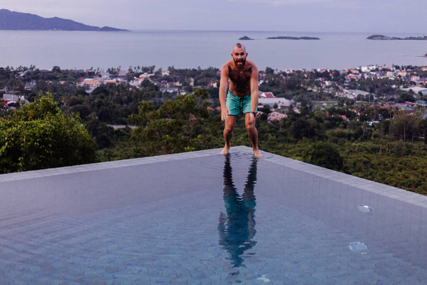 Hezký tetovaný běloch skáče v nekonečném bazénu v luxusní vile, tropický výhled na moře. - Fotografie, Obrázek