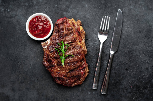 Grilovaný hovězí steak s příbory, kořením a bylinkami na tmavém dřevěném pozadí - Fotografie, Obrázek